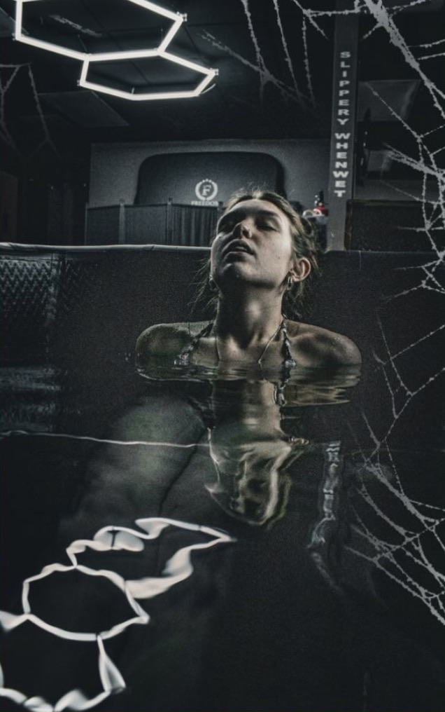 A guest leaning back in the cold plunge at Freedom, eyes closed, under the hexagon lights, with Halloween spiderwebs over the photo.